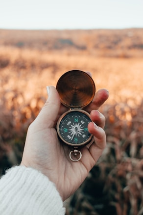 A hand is holding an open brass compass against a blurred natural background, suggesting a field or open landscape. The compass needle points ahead, and the person's sweater is visible, indicating it might be cool weather.