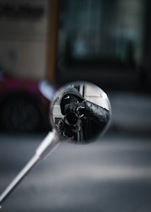 A reflection of a person holding a camera is captured in a round mirror attached to a metal rod. The person is wearing dark clothing, possibly a jacket with gloves, and appears to be looking through the camera lens. The background is blurred, focusing attention on the mirror's reflection.