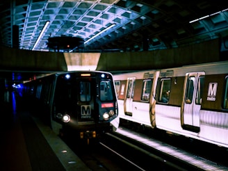 A subway train with the 'M metro' logo is stationed at an underground metro platform. The scene is dimly lit with artificial lights reflecting off the curved, metallic ceiling, creating an urban and somewhat futuristic atmosphere. The train cars are silver with dark windows, and there are few people visible inside.