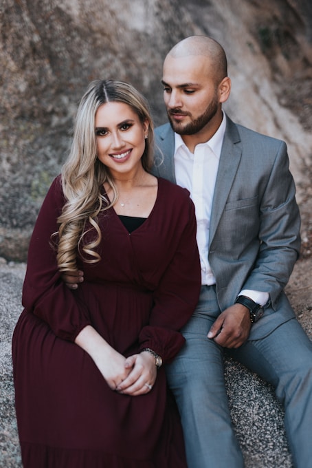 A couple is sitting close together on a rocky surface. The woman is wearing a long maroon dress and smiling at the camera. The man, dressed in a gray suit with a white shirt, looks at her warmly. Their body language suggests a close and affectionate relationship.