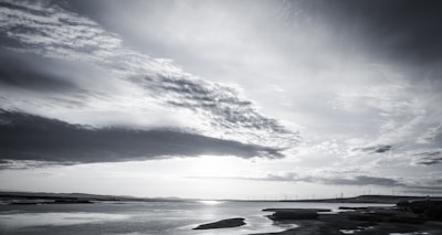 A scenic landscape featuring a wide expanse of water with a distant shoreline. The sky is overcast with dramatic clouds, creating a reflective effect on the water's surface. A line of wind turbines is visible on the horizon, suggesting an emphasis on clean energy. The overall atmosphere is tranquil and serene.