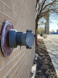 A close-up of a gray fire department connection on the side of a brick building. Around the connection is a slightly weathered red ring with some text. In the background, there is a sidewalk with some scattered mulch to the right, a few trees without leaves, and some distant objects that appear to be part of an urban or suburban street scene.