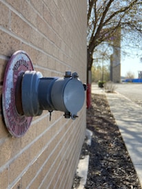 A close-up of a gray fire department connection on the side of a brick building. Around the connection is a slightly weathered red ring with some text. In the background, there is a sidewalk with some scattered mulch to the right, a few trees without leaves, and some distant objects that appear to be part of an urban or suburban street scene.