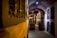 A dimly lit wine cellar with brick archways and metal gates lining the walls. Several large wooden barrels are positioned along the stone floor, creating a rustic atmosphere.
