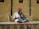 A musician sits cross-legged on a ledge, playing a traditional string instrument. He wears a colorful red turban and a white shirt, with a vibrant instrument resting against the wall. The wall behind him is textured and beige, casting shadows from the sunlight.