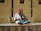A musician sits cross-legged on a ledge, playing a traditional string instrument. He wears a colorful red turban and a white shirt, with a vibrant instrument resting against the wall. The wall behind him is textured and beige, casting shadows from the sunlight.