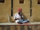 A musician sits cross-legged on a ledge, playing a traditional string instrument. He wears a colorful red turban and a white shirt, with a vibrant instrument resting against the wall. The wall behind him is textured and beige, casting shadows from the sunlight.