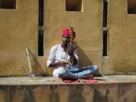A musician sits cross-legged on a ledge, playing a traditional string instrument. He wears a colorful red turban and a white shirt, with a vibrant instrument resting against the wall. The wall behind him is textured and beige, casting shadows from the sunlight.