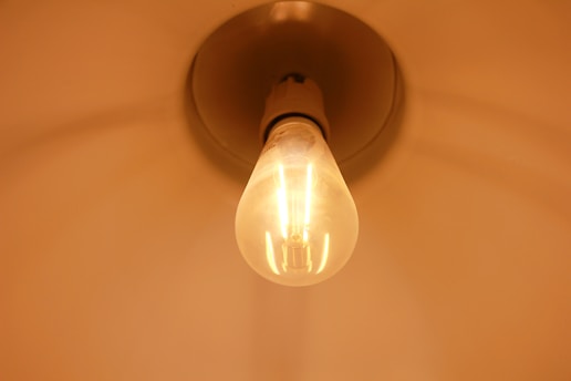 A close-up view of a lit incandescent light bulb, emitting a warm, gentle glow. The bulb is mounted in its socket against a plain background.