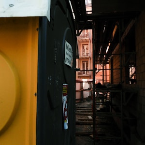 A construction site is visible with scaffolding along the side of a building. A yellow portable toilet stands on one side, featuring a sign with contact information. The construction area shows some building materials and a partially built structure, with a narrow view of windows from an adjacent building.