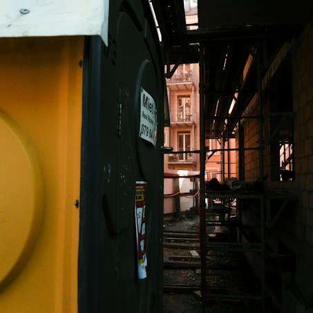 A construction site is visible with scaffolding along the side of a building. A yellow portable toilet stands on one side, featuring a sign with contact information. The construction area shows some building materials and a partially built structure, with a narrow view of windows from an adjacent building.