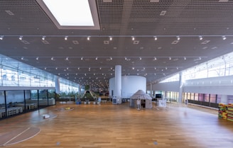 A spacious, modern indoor area with a large wooden floor and high ceilings. The ceiling features a grid pattern with numerous spotlights. On the left side, there are sections with glass walls, revealing plants and seating arrangements. In the center, a round white column supports the structure, and there are various installations, including a tent-like structure. The area is well-lit with natural light coming from large windows.