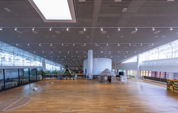 A spacious, modern indoor area with a large wooden floor and high ceilings. The ceiling features a grid pattern with numerous spotlights. On the left side, there are sections with glass walls, revealing plants and seating arrangements. In the center, a round white column supports the structure, and there are various installations, including a tent-like structure. The area is well-lit with natural light coming from large windows.