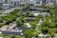 A rooftop garden featuring lush greenery with numerous trees and small glass structures. In the background, a cityscape with modern buildings and a curved structure with vibrant red, yellow, green, and blue panels can be seen.