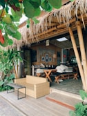 A cozy outdoor cafe with bamboo structures and a thatched roof. The seating area includes a wicker sofa and a wooden table with benches, surrounded by lush green plants. The decor features framed pictures and a chalkboard inside, adding to the rustic charm.