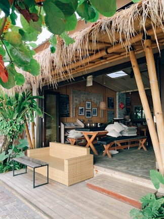 A cozy outdoor cafe with bamboo structures and a thatched roof. The seating area includes a wicker sofa and a wooden table with benches, surrounded by lush green plants. The decor features framed pictures and a chalkboard inside, adding to the rustic charm.