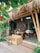 A cozy outdoor cafe with bamboo structures and a thatched roof. The seating area includes a wicker sofa and a wooden table with benches, surrounded by lush green plants. The decor features framed pictures and a chalkboard inside, adding to the rustic charm.