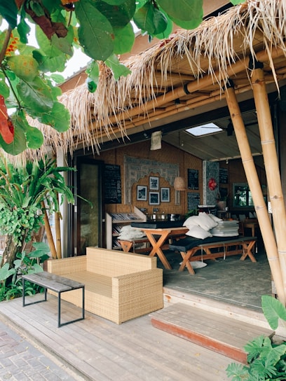 A cozy outdoor cafe with bamboo structures and a thatched roof. The seating area includes a wicker sofa and a wooden table with benches, surrounded by lush green plants. The decor features framed pictures and a chalkboard inside, adding to the rustic charm.