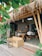A cozy outdoor cafe with bamboo structures and a thatched roof. The seating area includes a wicker sofa and a wooden table with benches, surrounded by lush green plants. The decor features framed pictures and a chalkboard inside, adding to the rustic charm.