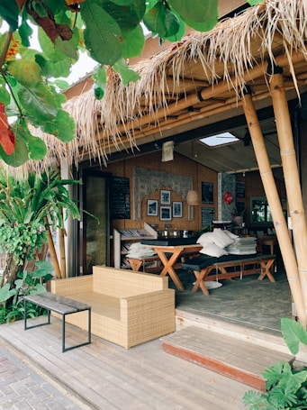 A cozy outdoor cafe with bamboo structures and a thatched roof. The seating area includes a wicker sofa and a wooden table with benches, surrounded by lush green plants. The decor features framed pictures and a chalkboard inside, adding to the rustic charm.