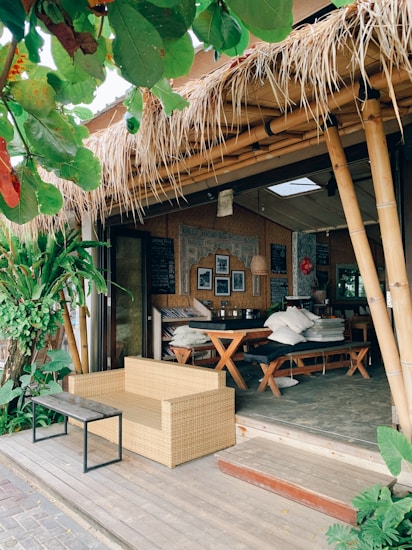 A cozy outdoor cafe with bamboo structures and a thatched roof. The seating area includes a wicker sofa and a wooden table with benches, surrounded by lush green plants. The decor features framed pictures and a chalkboard inside, adding to the rustic charm.