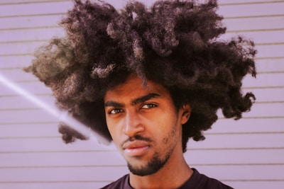 A person with a large, voluminous afro hairstyle stands in front of a light-colored, horizontally striped background. The lighting creates a soft glare in the bottom left corner, adding a unique effect to the image. The person has a contemplative expression.