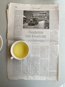 A newspaper is lying open on a table with a cup of light yellow beverage placed on the lower left side of the newspaper. The newspaper's headline reads 'Flexibilit&auml;t und Kreativit&auml;t', and it includes a black and white photograph of a vintage car beside a road sign. The text is printed in German.