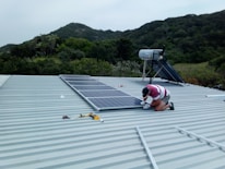 A person is installing solar panels on a metal rooftop with lush green hills in the background. The person is kneeling and using tools to secure the panels. The surrounding environment appears to be a natural, scenic area.