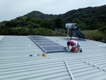 A person is installing solar panels on a metal rooftop with lush green hills in the background. The person is kneeling and using tools to secure the panels. The surrounding environment appears to be a natural, scenic area.