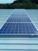 A solar panel installed on a metal roof with lush greenery in the background, under a clear sky.
