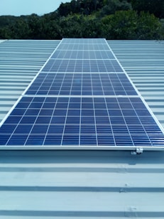 A solar panel installed on a metal roof with lush greenery in the background, under a clear sky.
