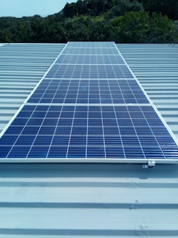 A solar panel installed on a metal roof with lush greenery in the background, under a clear sky.