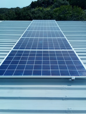 A solar panel installed on a metal roof with lush greenery in the background, under a clear sky.