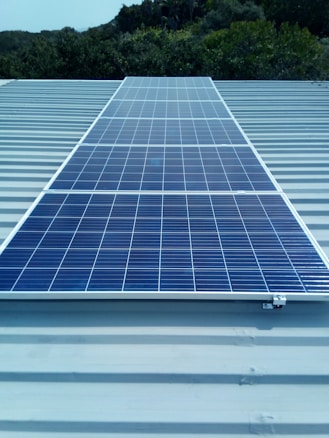 A solar panel installed on a metal roof with lush greenery in the background, under a clear sky.