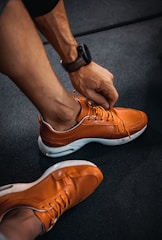 A person is tying the laces of a bright orange sneaker with a perforated design. The shoes have white soles, and the individual is wearing a dark wristwatch. The setting appears to be an indoor floor with a dark, textured surface.