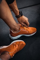 A person is tying the laces of a bright orange sneaker with a perforated design. The shoes have white soles, and the individual is wearing a dark wristwatch. The setting appears to be an indoor floor with a dark, textured surface.