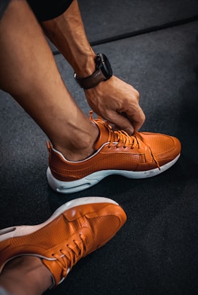 A person is tying the laces of a bright orange sneaker with a perforated design. The shoes have white soles, and the individual is wearing a dark wristwatch. The setting appears to be an indoor floor with a dark, textured surface.