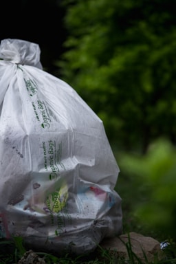 A large white bag labeled 'Organic Bags' is filled with various trash items, resting on the ground surrounded by lush green vegetation, creating a juxtaposition between nature and waste.