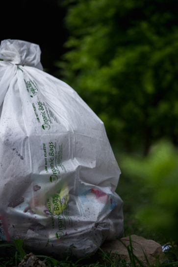 A large white bag labeled 'Organic Bags' is filled with various trash items, resting on the ground surrounded by lush green vegetation, creating a juxtaposition between nature and waste.