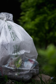 A large white bag labeled 'Organic Bags' is filled with various trash items, resting on the ground surrounded by lush green vegetation, creating a juxtaposition between nature and waste.