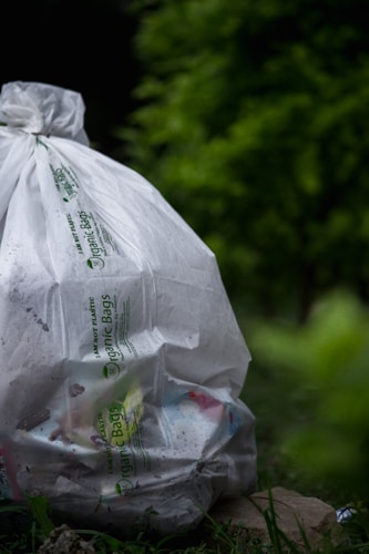 A large white bag labeled 'Organic Bags' is filled with various trash items, resting on the ground surrounded by lush green vegetation, creating a juxtaposition between nature and waste.