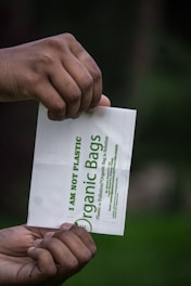 Two hands are holding a small white bag with green text that reads 'I am not plastic' and 'Organic Bags'. The background is blurred, showcasing a dark green hue, possibly indicating a natural setting.