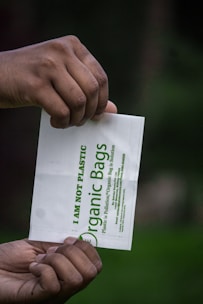 Two hands are holding a small white bag with green text that reads 'I am not plastic' and 'Organic Bags'. The background is blurred, showcasing a dark green hue, possibly indicating a natural setting.