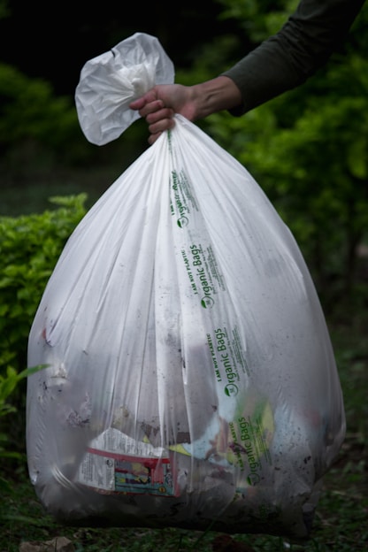 A hand holds a large white plastic garbage bag filled with various items, visible through the semi-transparent material. The bag features printed text indicating it is an organic bag. The background consists of a blurred, green, leafy environment, suggesting an outdoor setting.