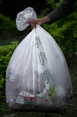 A hand holds a large white plastic garbage bag filled with various items, visible through the semi-transparent material. The bag features printed text indicating it is an organic bag. The background consists of a blurred, green, leafy environment, suggesting an outdoor setting.