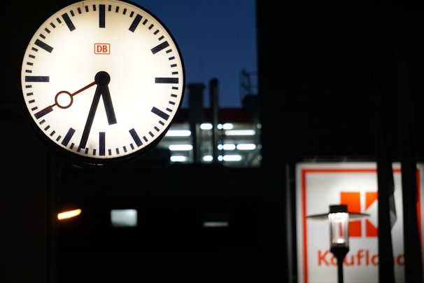 A large round clock with a white face and black markings is prominently displayed. It shows the time, with black hour and minute hands, and a red second hand. In the background, there's a blurry sign with red text and logo on a white background, possibly indicating a store or commercial building.