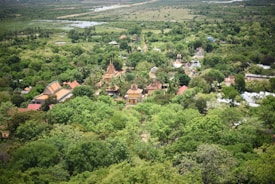 An expansive aerial view of a lush, green landscape with a collection of traditional architectural structures, including temples nestled among dense trees. The horizon shows a mix of fields and distant water bodies. The prominent presence of vegetation interspersed with colorful rooftops creates a harmonious blend with nature.