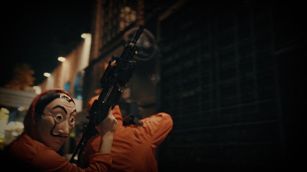 A person wearing a Salvador Dalí mask and a red outfit is holding a gun in a dimly lit urban setting. Another figure in similar clothing is visible in the background, creating a tense and mysterious atmosphere.