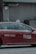 A red taxi with the branding 'Transcab' and a phone number is seen driving along a street. The vehicle is in motion, with a driver visible inside. The surroundings include a textured, gray stone building facade with some surveillance cameras mounted above.