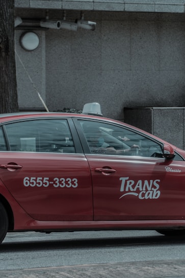 A red taxi with the branding 'Transcab' and a phone number is seen driving along a street. The vehicle is in motion, with a driver visible inside. The surroundings include a textured, gray stone building facade with some surveillance cameras mounted above.